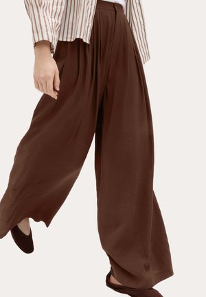 Persona con pantalones marrones anchos de talle alto y sueltos, combinados con una camiseta de rayas de manga larga y zapatos planos oscuros.