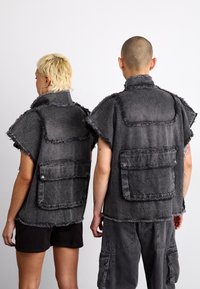 Γκρι τζιν γιλέκα με φθαρμένα άκρα, κοντά μανίκια και μεγάλα μπροστινά τσεπάκια. Υφασμάτινη επιφάνεια με υφή; διαθέτει εμφανή ραφή και κουμπώματα.