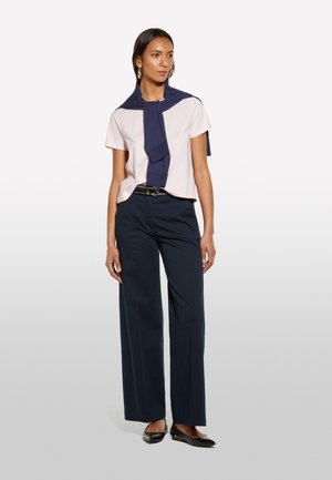 T-shirt rose clair, pull bleu marine noué autour des épaules, pantalon large bleu marine, ballerines noires, avec des boucles d'oreilles créoles.
