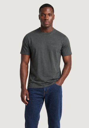 Uomo che indossa una maglietta a maniche corte grigio scuro e jeans blu, in piedi con una mano in tasca davanti a uno sfondo semplice.