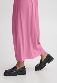 Mocasines negros con suelas de goma gruesa, combinados con un vestido largo rosa fluido, que muestra una tela suave y una abertura lateral.