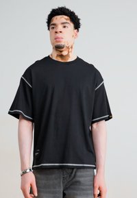 T-shirt noir à manches courtes avec des coutures blanches contrastées le long des ourlets, présentant une coupe décontractée et une texture de tissu douce.