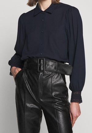 Kobieta w ciemnogranatowej bluzce z koronkowymi mankietami i bufkami na rękawach, włożona w czarne skórzane spodnie z wysokim stanem i szerokim paskiem.