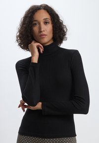 Haut à col roulé noir côtelé avec des manches longues, texture lisse, design ajusté et col haut. Porté avec un bas à motifs.
