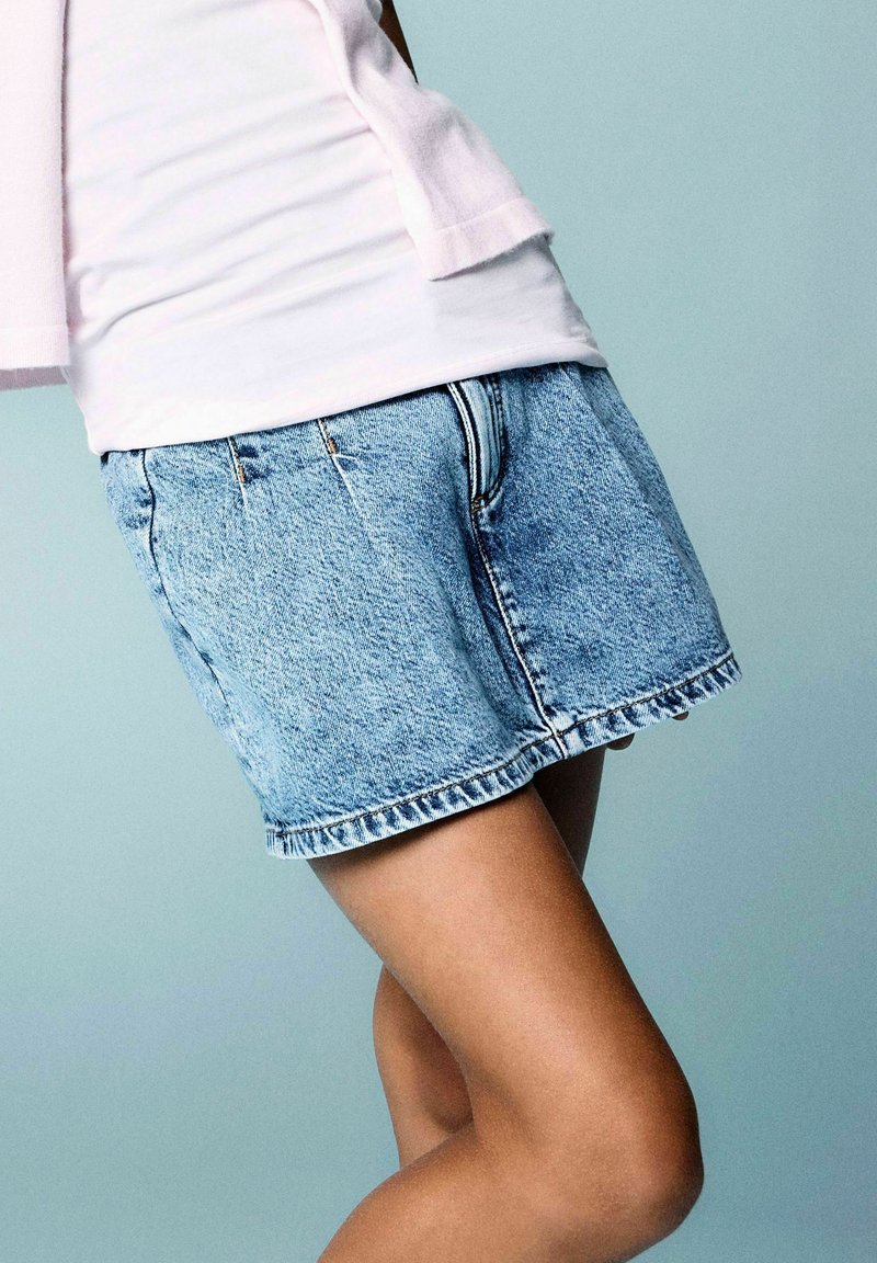 Personne portant un short en jean bleu et une chemise blanche debout devant un fond uni bleu clair.