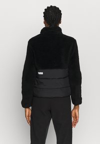 Zwarte jas met een gestructureerde fleece bovenkant en een gladde, gewatteerde onderkant. Heeft een opstaande kraag en een logo-detail op de achterkant.