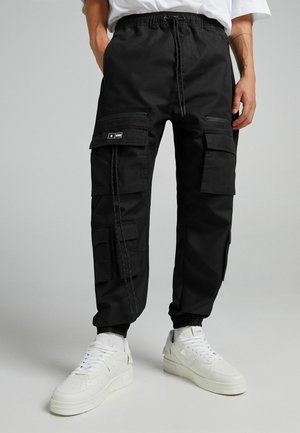 Homme portant un pantalon cargo noir avec plusieurs poches et des baskets blanches, debout devant un fond gris uni.