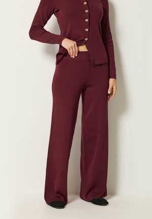 Personne portant un haut bordeaux à manches longues boutonné et un pantalon large assorti, debout devant un mur blanc, avec des chaussures noires visibles.