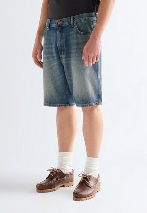 Mann iført falmede blå denimshorts, hvite crew-sokker og brune skinnbåtsko, stående mot en ensfarget bakgrunn.