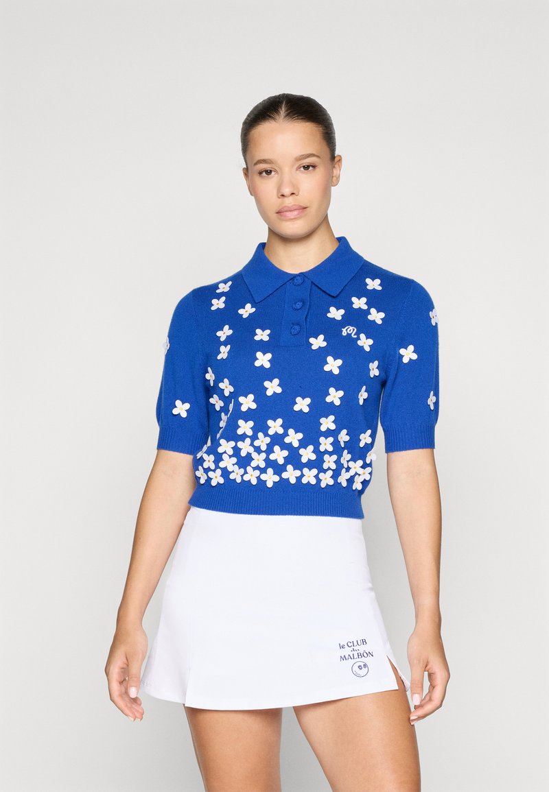 Blå strikket polo med korte ermer, med hvit blomsterbroderi og tre knapper ved kragen. Akkompagnert av et hvitt skjørt.