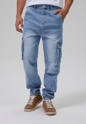 Mann trägt hellblaue Denim-Cargohose mit Seitentaschen und braune Wildleder-Sneakers mit weißen Schnürsenkeln, Hand in der Tasche, steht auf grauem Boden.