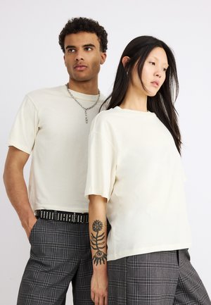 Δύο μοντέλα φορούν λευκά φαρδιά μπλουζάκια και γκρι καρό παντελόνια, ένα με τατουάζ ήλιου και λουλουδιού στο χέρι.