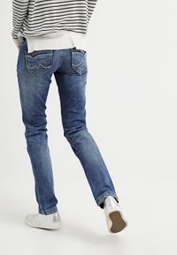 Jeans de mezclilla azul con un ajuste slim, un ligero desgastado y bolsillos traseros distintivos con costura. Combinados con zapatillas blancas y un suéter a rayas.