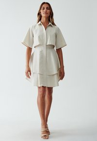 Crème-kleurige jurk met korte mouwen, kraag, gelaagd rokontwerp en een sluiting met knopen aan de voorkant. Gladde textuur met een minimalistische stijl.