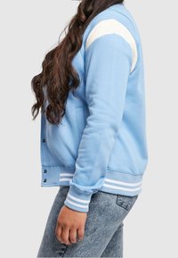 Person med långt mörkt hår som bär en ljusblå och vit collegejacka och ljustvättade jeans, sedd från sidan, midjan upp.