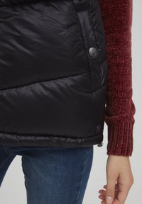 Crni puffer prsluk s glatkom, sjajnom teksturom, s patentnim zatvaranjem, gumbima na strani, i kontrastnim burgundskim rukavom.