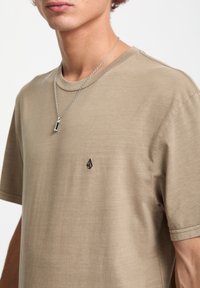 Beige bomulds T-shirt med rund halsudskæring og korte ærmer, med et lille sort logo på brystet. Sølv kæde halskæde synligt.