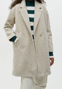 Manteau beige en mélange de laine avec col cranté, fermeture à double bouton et poches latérales. Porté par-dessus un pull rayé vert et blanc.