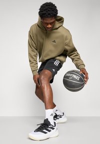Green zip-up hoodie, black shorts with three white stripes, white athletic shoes with black accents, holding a black basketball.
