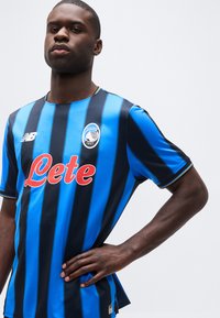 Jaunas vyras dėvi mėlynos ir juodos juostuotos Atalantos futbolo marškinėlius su raudonu „Lete“ rėmėjo logotipu, stovintis su viena ranka ant juosmens.