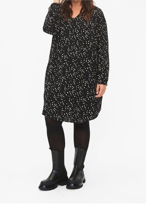 Robe noire à manches longues avec des points blancs éparpillés, dotée d'un décolleté en V et d'une coupe décontractée, associée à des bottines noires.