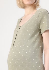 Grūtniecības kleita gaiši zaļā audumā ar V veida kakla izgriezumu, īsām piedurknēm un baltu zvaigžņu rakstu. Priekšpusē ir pogu detaļa.