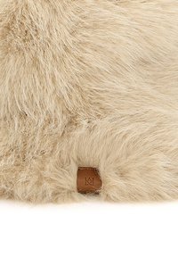 Beige faux fur materiale med en blød, fluffy tekstur, med et lille brunt lædermærke præget med initialer.