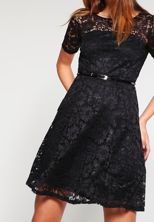 Frau trägt ein knielanges schwarzes Spitzenkleid mit kurzen Ärmeln und einem dünnen schwarzen Gürtel in der Taille.