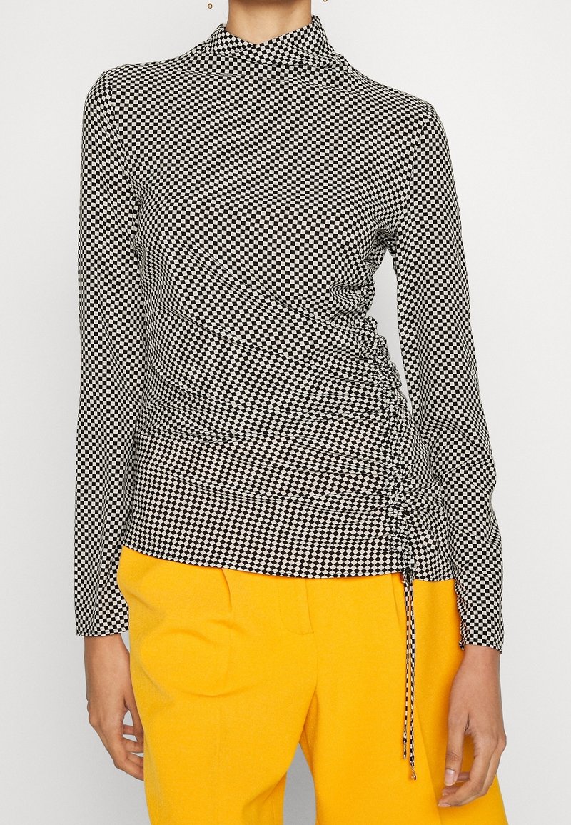 Černobílé kostkované dlouhé tričko s nařaseným detailem po straně a límečkem, doplněné žlutými kalhotami.