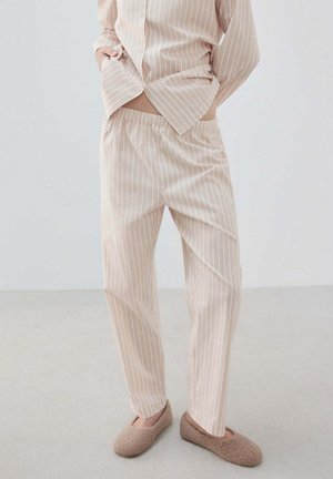 Personne portant un pyjama beige et blanc rayé et des chaussons marron pelucheux, debout les mains dans les poches sur un fond clair uni.