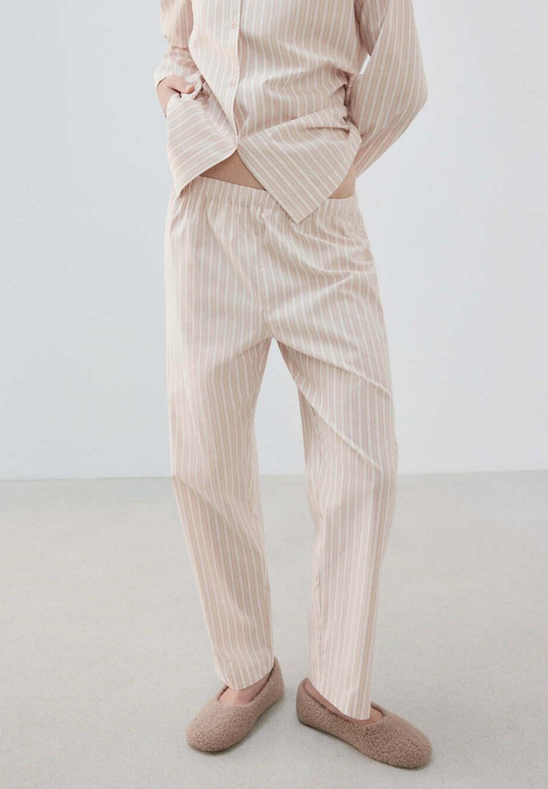 Personne portant un pyjama beige et blanc rayé et des chaussons marron pelucheux, debout les mains dans les poches sur un fond clair uni.