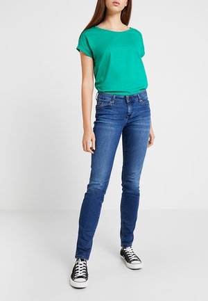 Woman wearing a green short-sleeve shirt, blue slim-fit jeans, and black low-top sneakers standing against a white background.