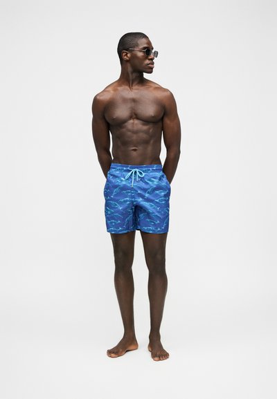 Άνδρας χωρίς πουκάμισο που φορά μπλε μαγιό με σχέδια καρχαρία και γυαλιά ηλίου, όρθιος ξυπόλητος μπροστά σε απλό ανοιχτό φόντο.