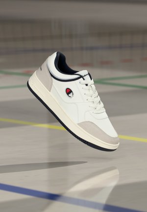 Hvid sneaker med ruskindsdetaljer, der har en rund tå, snørebånd foran og en sort gummisål. Broderet logo på siden.