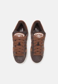 Bruna mockasneakers med bruna snören och tre diagonala ränder. Tungans omslag har en cirkulär logotyp med varumärkesnamnet i vitt.