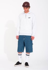 Sudadera de forro polar gris claro con cremallera y un pequeño logo, combinada con pantalones cortos de mezclilla azules hasta la rodilla, calcetines blancos con rayas negras y zapatillas negras.