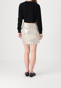 Femme portant un haut noir à manches longues et une mini-jupe à franges argentées en sequins, debout de dos à la caméra sur un fond blanc uni.