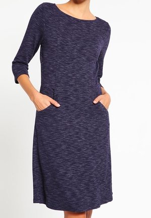 Mujer con un vestido azul oscuro texturizado hasta la rodilla, de manga tres cuartos y con las manos en los bolsillos laterales.