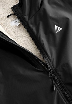 Crna jakna s patentnim zatvaračem sprijeda i flis podlogom. Uključuje bijelo brendiranje i teksturiranu vanjštinu koja pruža toplinu i izdržljivost.