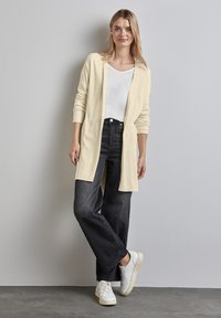 Długi kardigan z otwartym przodem w jasnym kremowym kolorze, założony na biały t-shirt. Połączony z czarnymi jeansami o szerokich nogawkach i białymi trampkami. Gładka tekstura.