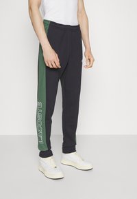 Navy sweatpants with green side panel, elastic waistband, and white "LACOSTE" logo. Made of soft fabric, ideal for casual wear.