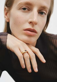 Anillo de oro con una piedra preciosa ovalada púrpura en el centro, llevado en una mano con esmalte de uñas natural, sobre un fondo neutro.