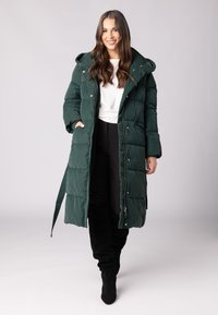 Lang, donkergroen pufferjas met capuchon, met een gequilt ontwerp, vooraan met rits en drukknoopsluiting, gecombineerd met zwarte slim fit broek en laarzen.