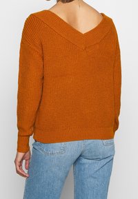 Strickpullover in verbranntem Orange mit V-Ausschnitt, geripptem Kragen und Bündchen. Strukturierter Stoff mit lockerer Passform, kombiniert mit hellblauen Jeans.