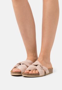 Ljust rosa mocka slidesandal med knutdesign, platt korksula och öppen tå. Slät textur och avslappnad, minimalistisk stil.