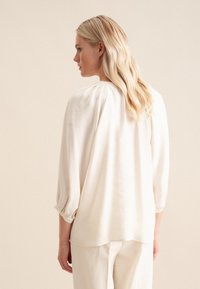 Lys beige bluse med trekvartærmer, blød tekstur, løs pasform, sammensamlet detalje ved halsen og en lige kant, parret med matchende bukser.