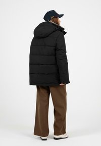 Černá péřová bunda s kapucí, s horizontálně prošívanými sekcemi. V kombinaci s hnědými kalhotami širokého střihu a bílými botami.