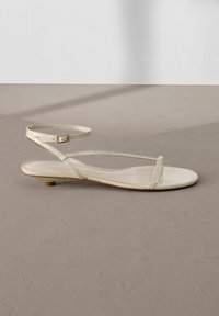 Sandale plate beige avec fines lanières, boucle à la cheville et petit talon sur une surface texturée marron clair.