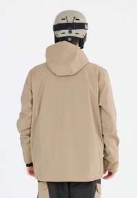 Beige ytterjacka med huva, tillverkad av slätt, lätt material. Bärs med en beige skidhuvudskydd, som visar rena linjer och minimalistisk design.