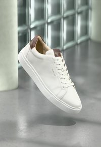 Adidași din piele albă cu talpă de călcâi și limbă maro, logo-ul "GANT" pe lateral și pe limbă, plutind deasupra unei podele lucioase de gri și a unui Zid din blocuri de sticlă.